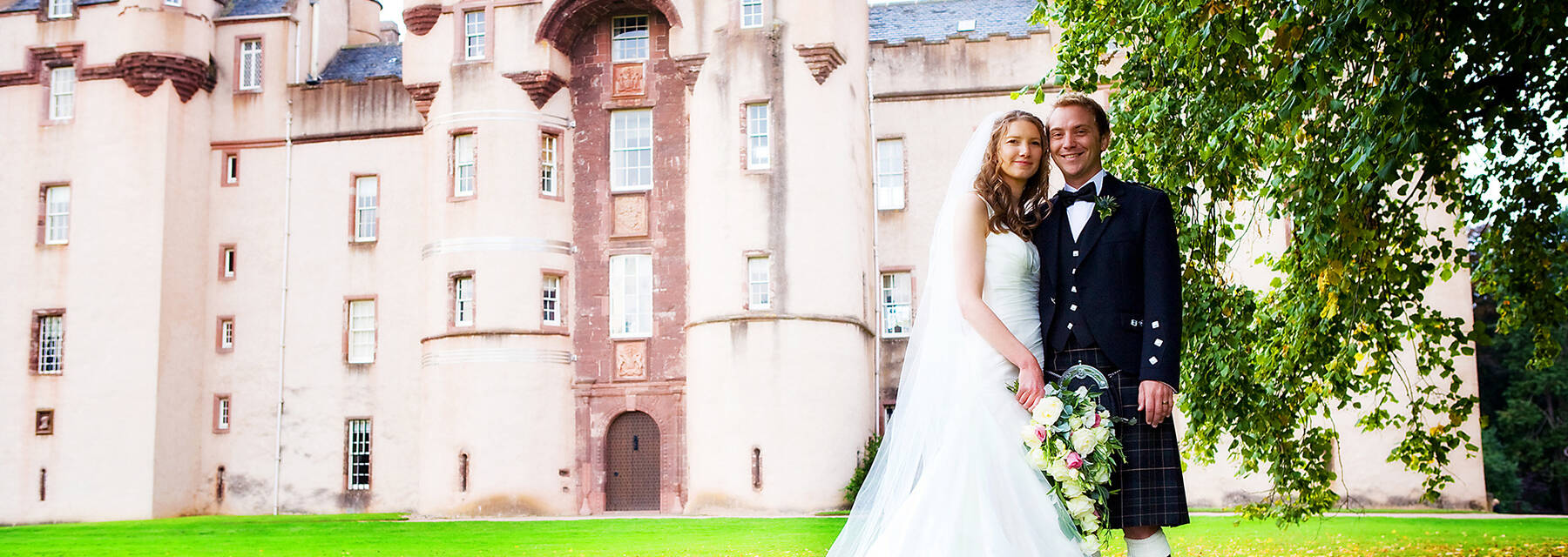 A wedding couple stand on the lawn in front of Fyvie Castle, beside a large tree.