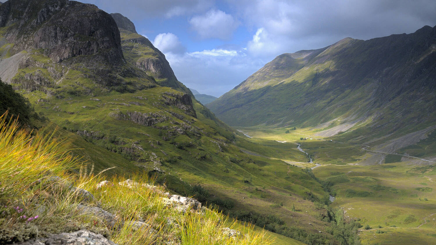 A view looking down the glen of Glencoe on a sunny day, with the steep mountains on either side casting shadows on the opposite side.