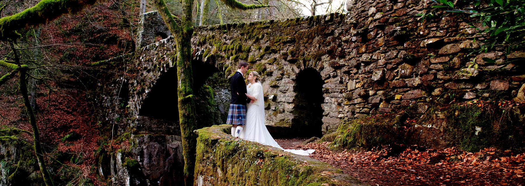 Wedding couple standing by the stone bridge at the Hermitage. Moss-covered branches and old stonework contrast to the rich orange and red autumn leaves on the path and trees.