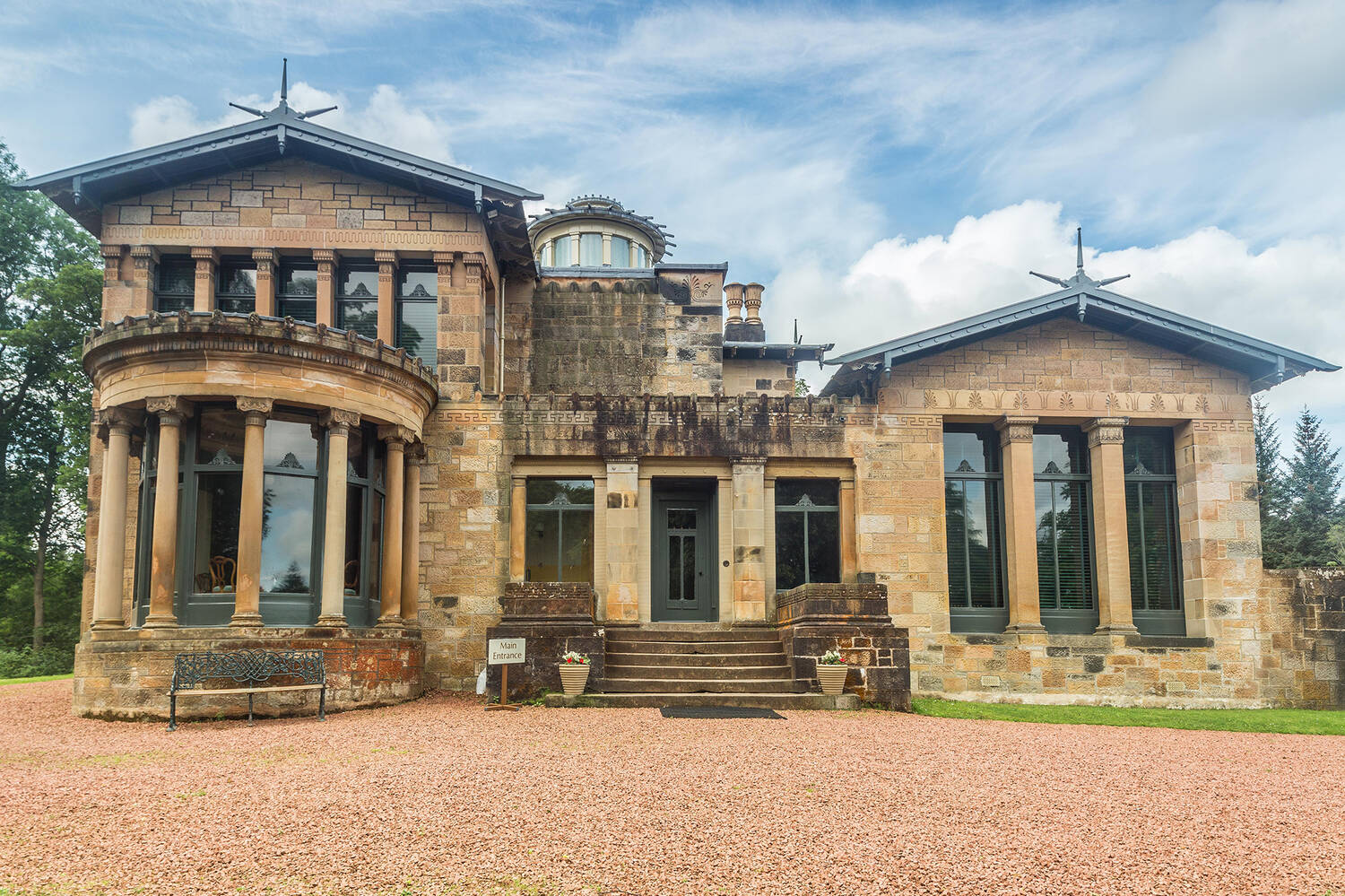A view of the grand front exterior of Holmwood. It is a sandstone building, with columns and classical motifs. A large gravel area lies in front of the house.