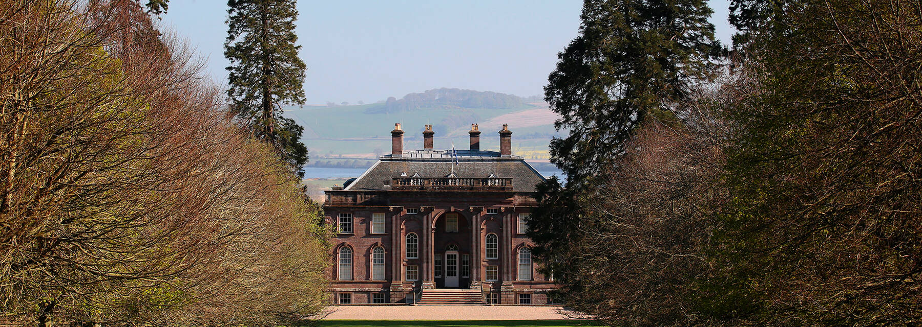 A view of a grand Georgian country house, seen from a wide grassy avenue of beech trees. Hills can be seen across the estuary in the background.