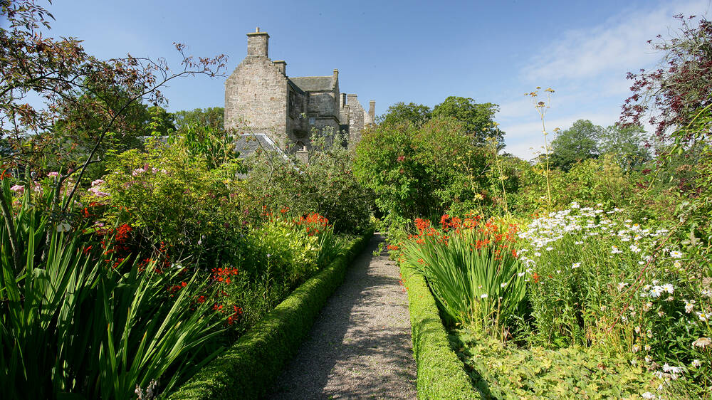 A view of the side of Kellie Castle from the very lush and colourful garden. A narrow gravel path, lined by small box hedges, leads towards the castle, between beds of white and red flowers. The sky is blue.