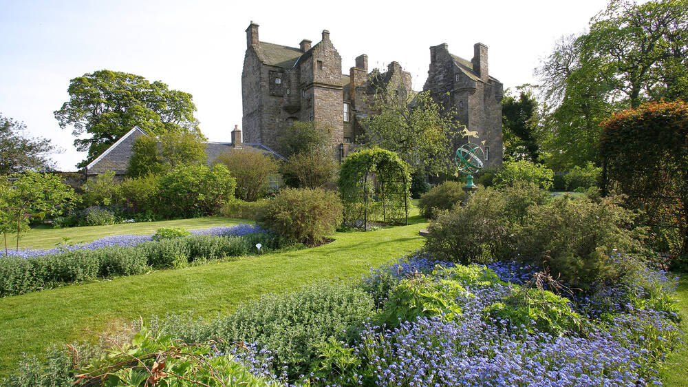 A view of Kellie Castle with its towers and chimneys, seen from a colourful garden. The bed in the foreground is filled with small blue flowers, mirroring the sky!