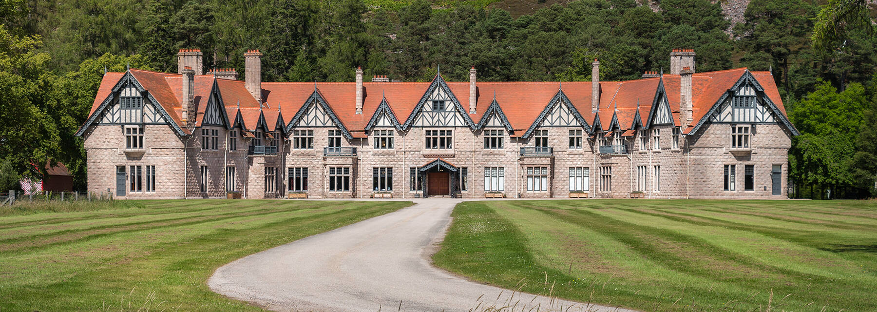 A long and grand two-storey lodge building, with a red-tiled roof, stands with an enormous lawn in front. Behind rises the steep slopes of a heather-covered mountain.