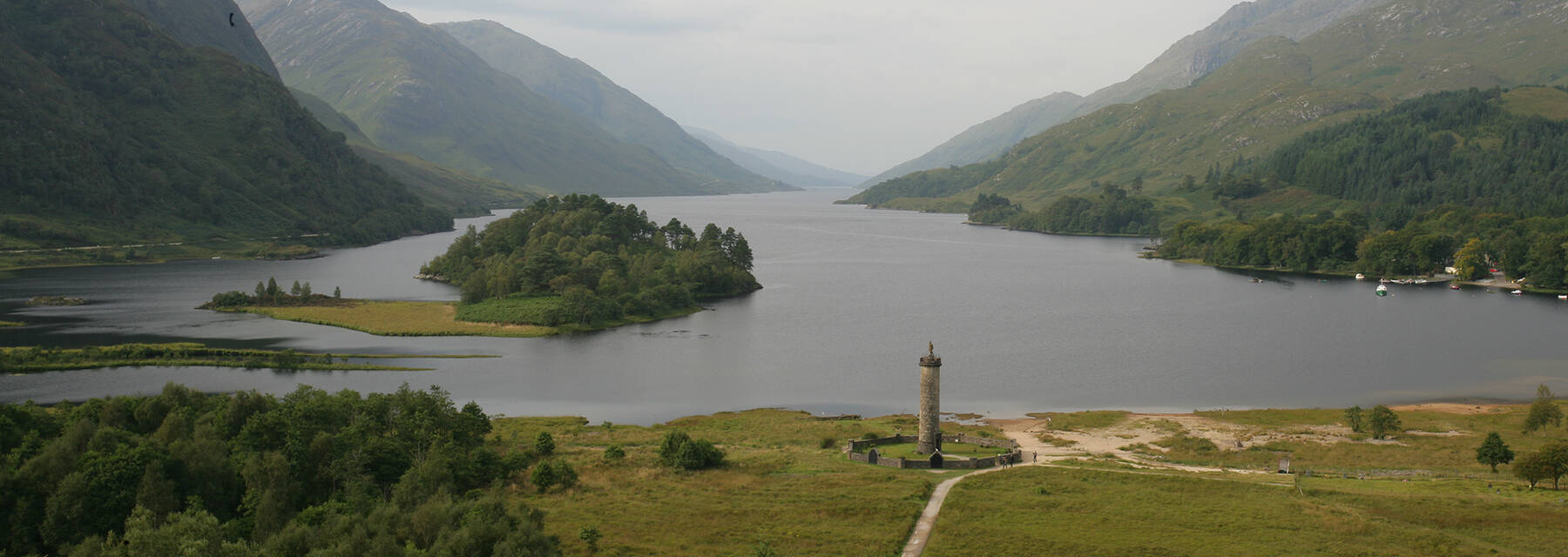 An aerial view of Glenfinnan from the viaduct area, looking down towards the monument at the edge of the loch. The loch and mountains stretch away into the distance.