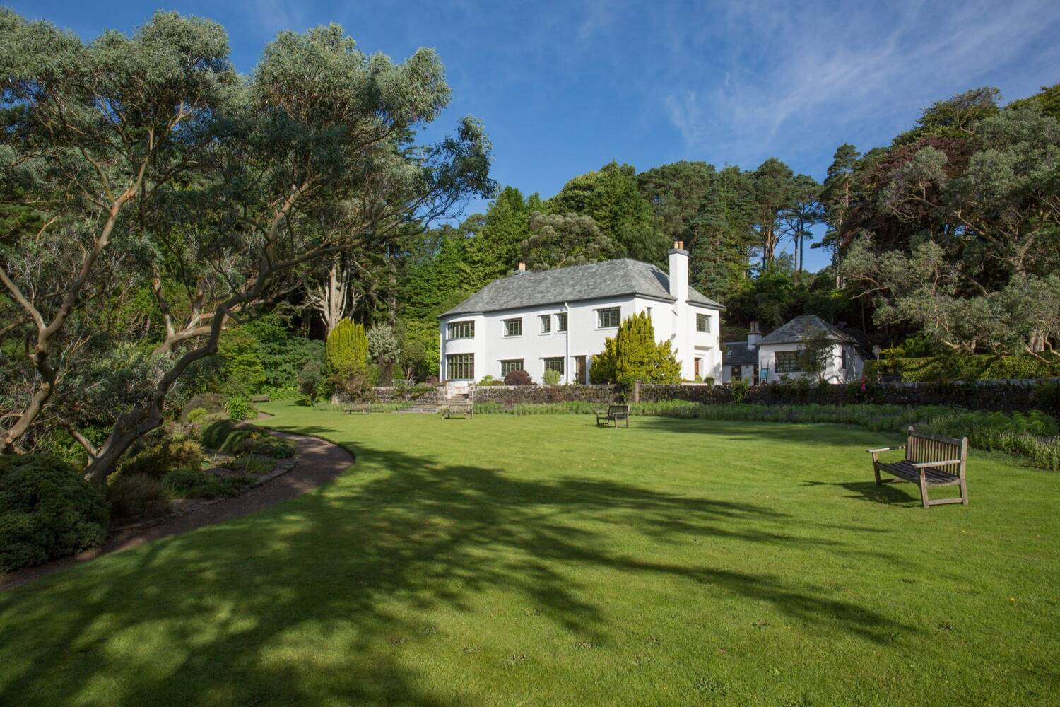 A view of the white stone Inverewe House, sitting at the centre of a large garden. A large lawn area is in the foreground surrounded by tall trees. A wooden garden bench sits in the middle of the lawn.
