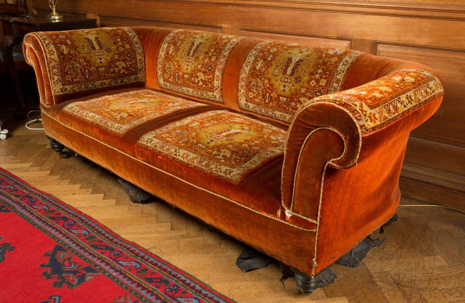 Sofa in Cedar Room, Pollok House. It's a three seater and covered with a Turkish rug-type fabric in bold orange colours. It stands on a wooden floor against the cedar-panelled wall.