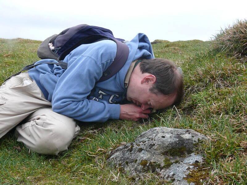 A man lies on a grassy hillside with his face almost resting on the grass. He is looking at a plant through a small lens.