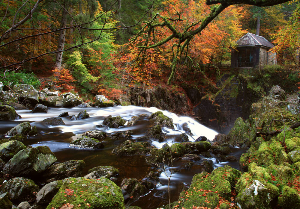 A fast-flowing river with large boulders in it, with trees in autumn colours on the bank. There is a round building in the background overlooking a waterfall.