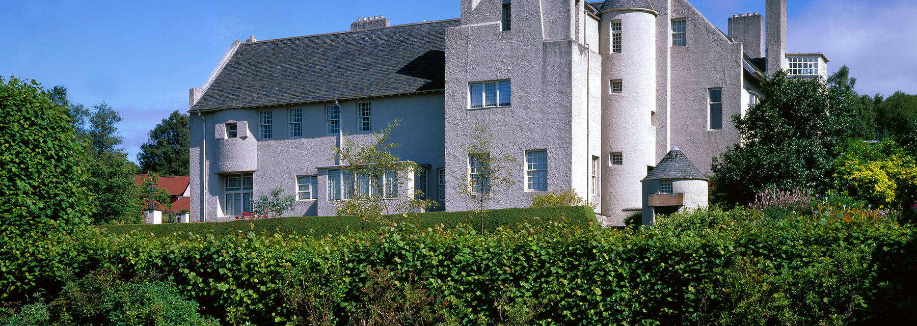 An exterior view of the Hill House on a sunny day, from the garden below. The white cement-harled walls and grey slate roof tiles stand out against the bright blue sky.