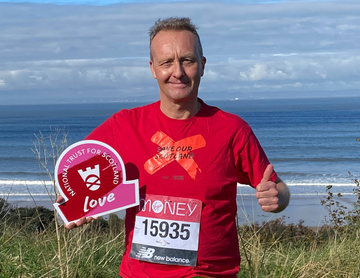 A man in a bright red t-shirt with a runner's number pinned to the front stands on a grassy area in front of the sea. In one hand he holds an omega-shaped board that says Love; he's giving a thumbs up sign with his other hand.