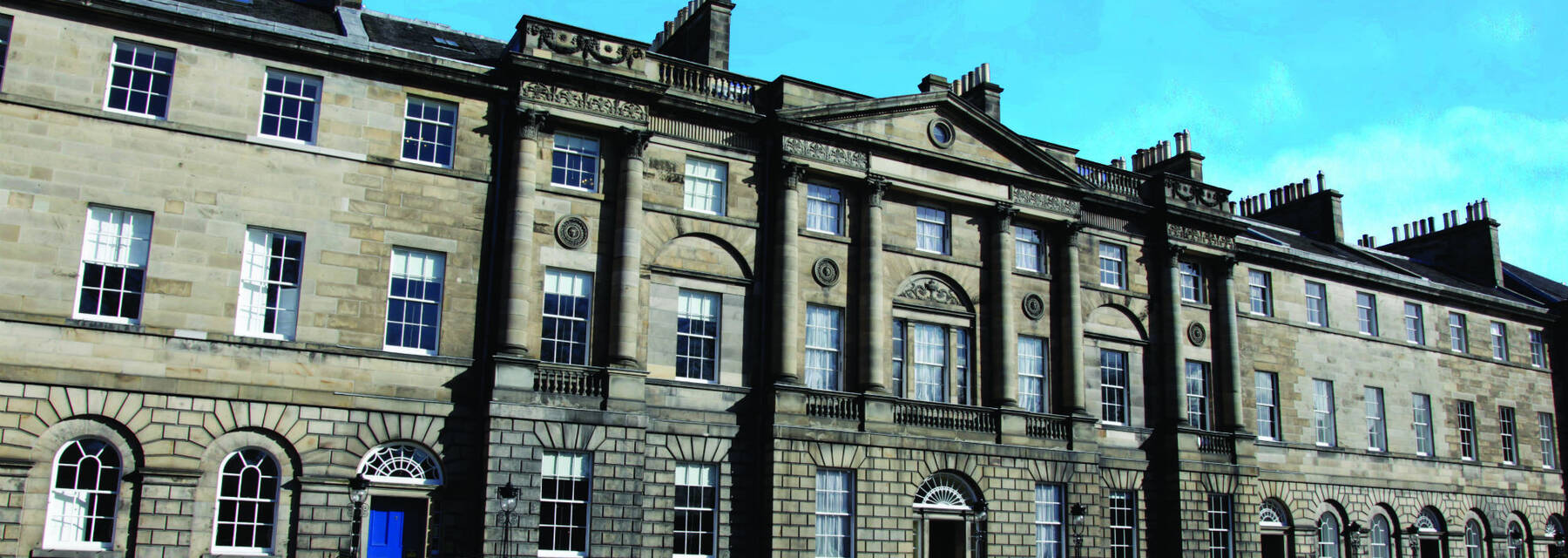 A view of the frontage of the Georgian House and neighbouring Robert Adam-designed buildings, in Charlotte Square. The Georgian House has a blue front door in this image, mirroring the blue sky overhead.