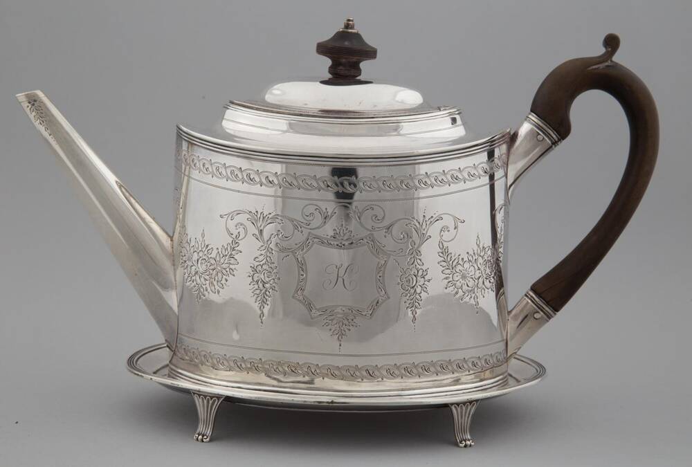 Silver teapot with a dark handle and knob on the top. It is sitting on a silver stand.