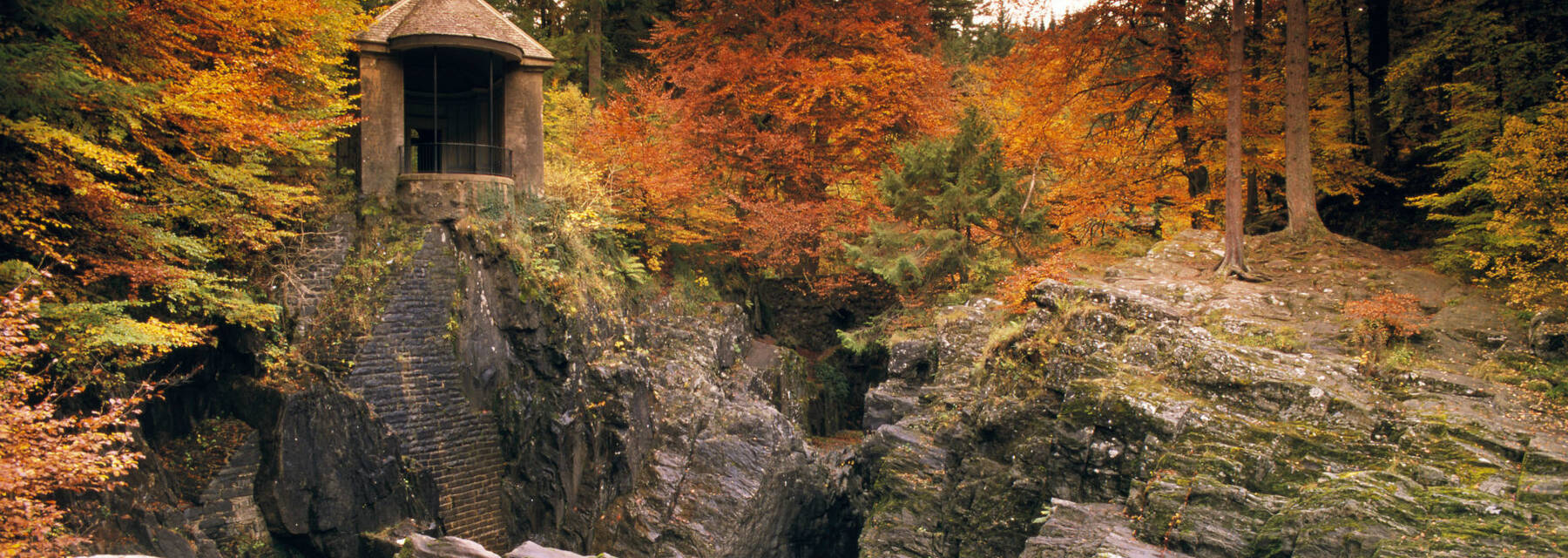 A folly stands on a rocky crag, overlooking a river falling over a waterfall. In the background is a forest bursting with rich autumn colour.
