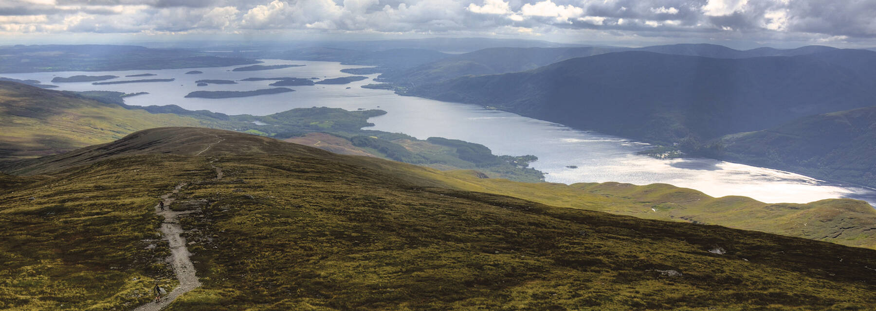 A view of Loch Lomond from Ben Lomond. The sun shines through dark clouds and shimmers on the water. The main mountain path can also be seen snaking across the open hillside.