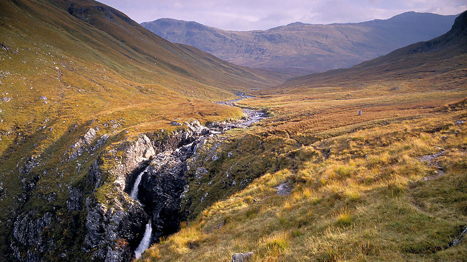 A river runs through a moorland valley, surrounded by tall mountains. In the foreground, it plunges over a long rocky drop, creating a white waterfall.