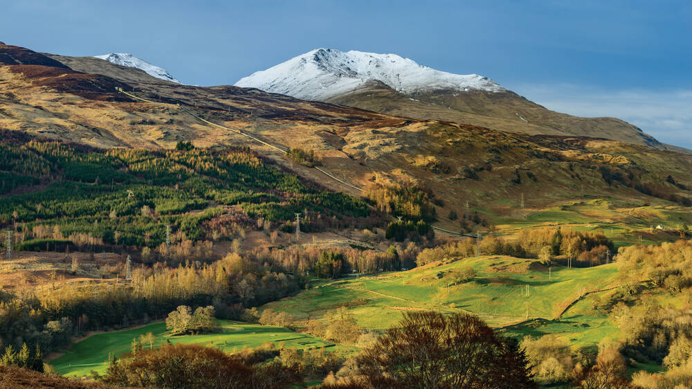 The snow-capped peak of Ben Lawers rises, with grassy foothills in the foreground. The sky is bright blue.