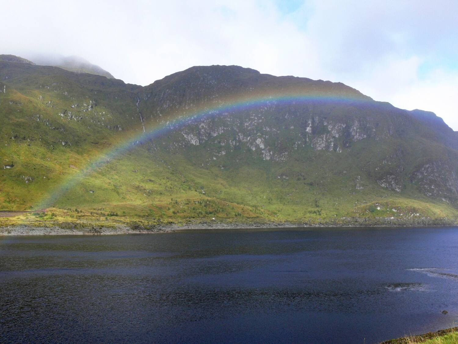 A low rainbow arches across a loch, with mountains behind.