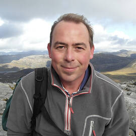 A photo of the upper half of a man out on a mountain. He has short brown hair and is wearing a grey fleece jacket with a rucksack.