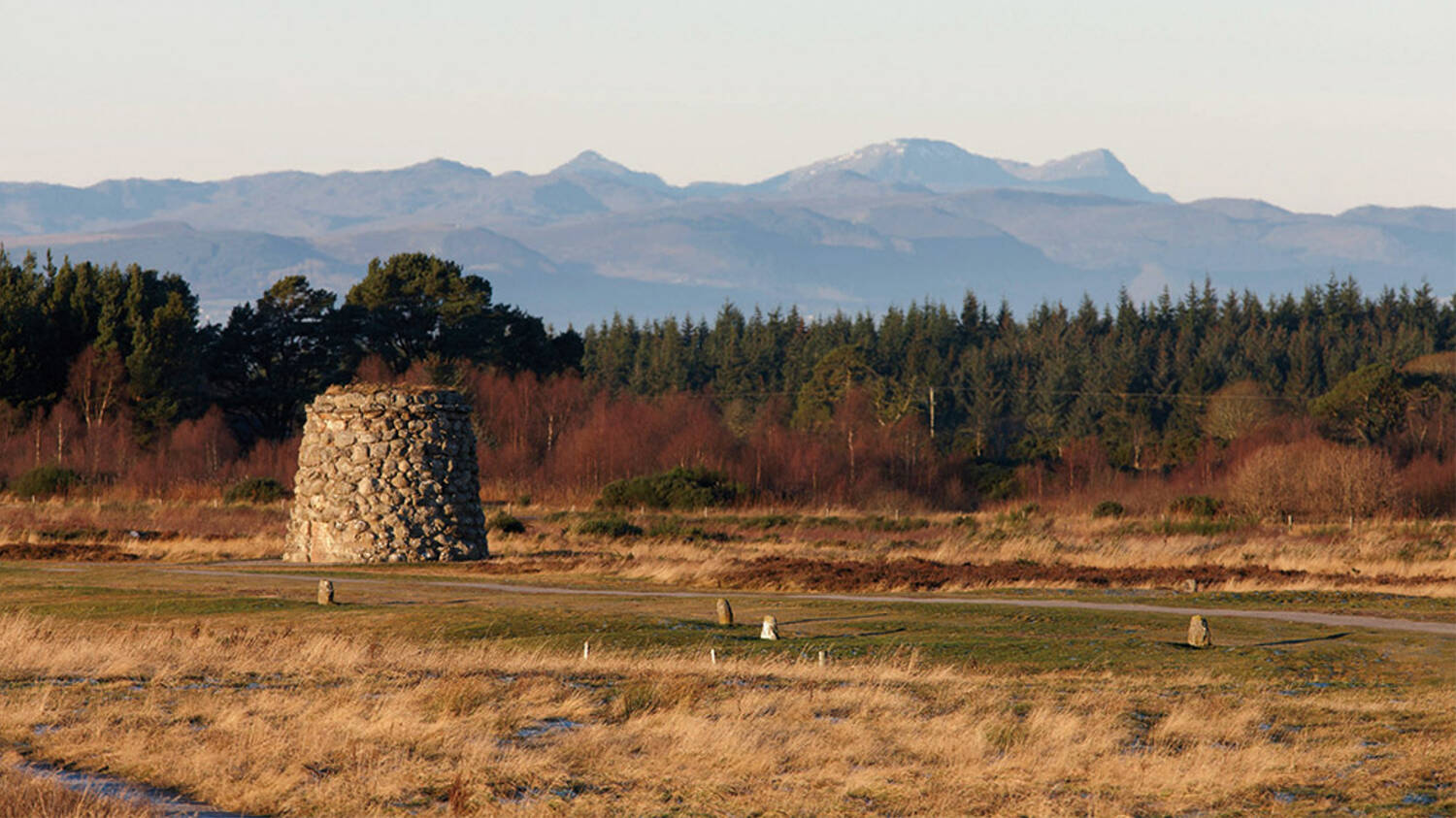A large round stone cairn stands in the middle of a battlefield. Mountains and forests can be seen in the distance.