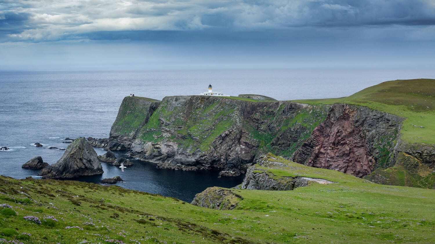 A white lighthouse stands at the edge of green cliffs on a headland. The sky overhead is heavy and grey, with occasional shafts of sunlight shining through.