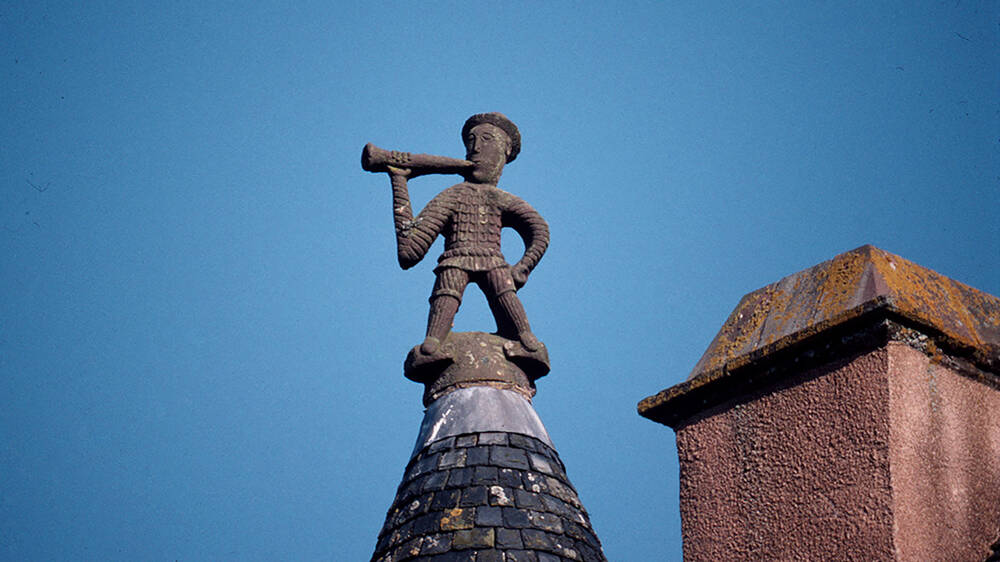 A stone carving of a man blowing a hunting horn perches on top of a castle turret, beside a pink-harled stone chimney.