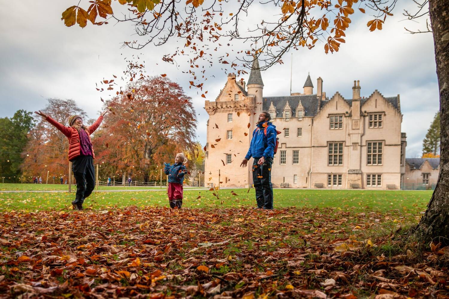 A family is playing among fallen autumn leaves underneath a tree. A grand castle is behind them.