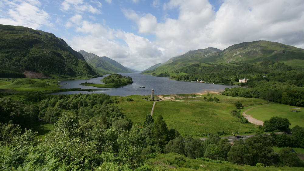 An aerial view of Glenfinnan Monument, taken from above the viaduct and looking towards the loch and mountains in the background.