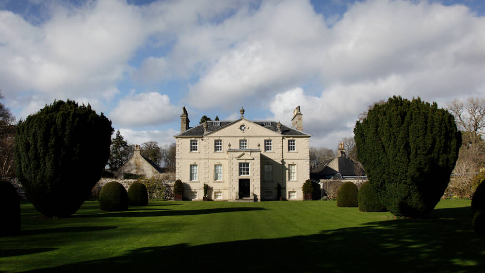 A symmetrical, Georgian-style large country house stands at the top of an immaculate lawn, with large yew trees to either side. It is a sunny day with fluffy white clouds across a blue sky.