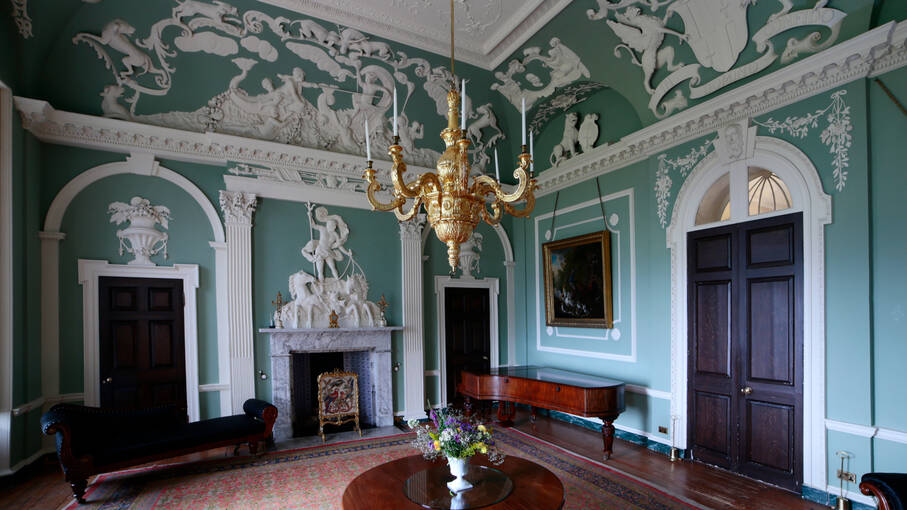 A drawing room with mint green walls and elaborate white plasterwork decoration. A gold chandelier hangs in the centre of the room above a round polished table.