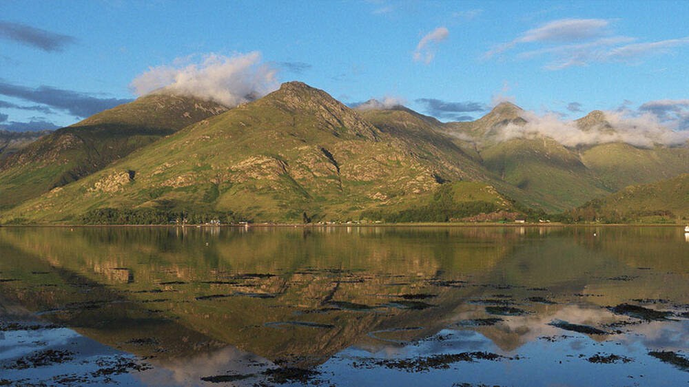 Towering mountains are reflected in the still clear waters of a loch.
