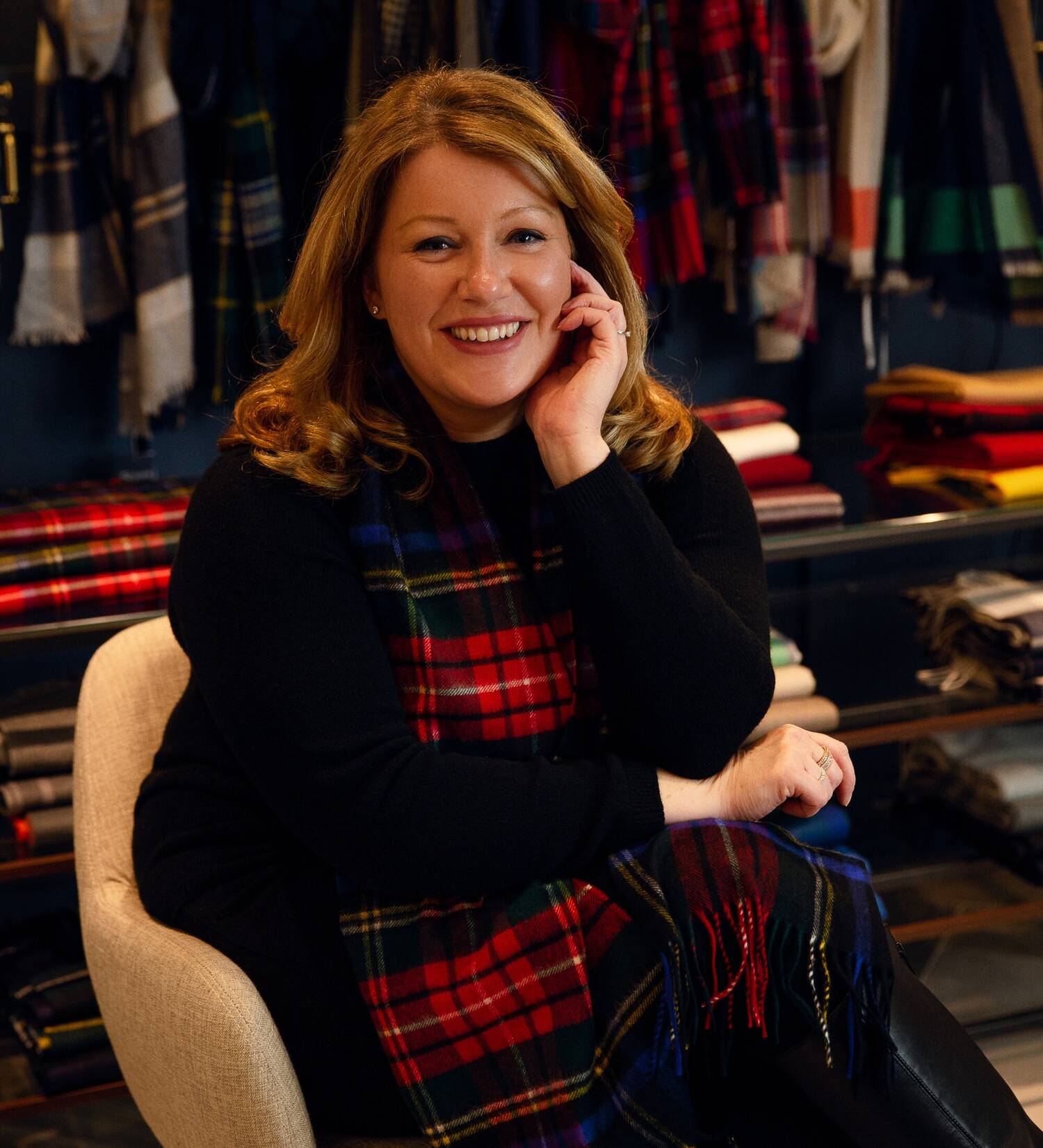 A smiling woman is sitting on a chair, wearing a tartan shawl.