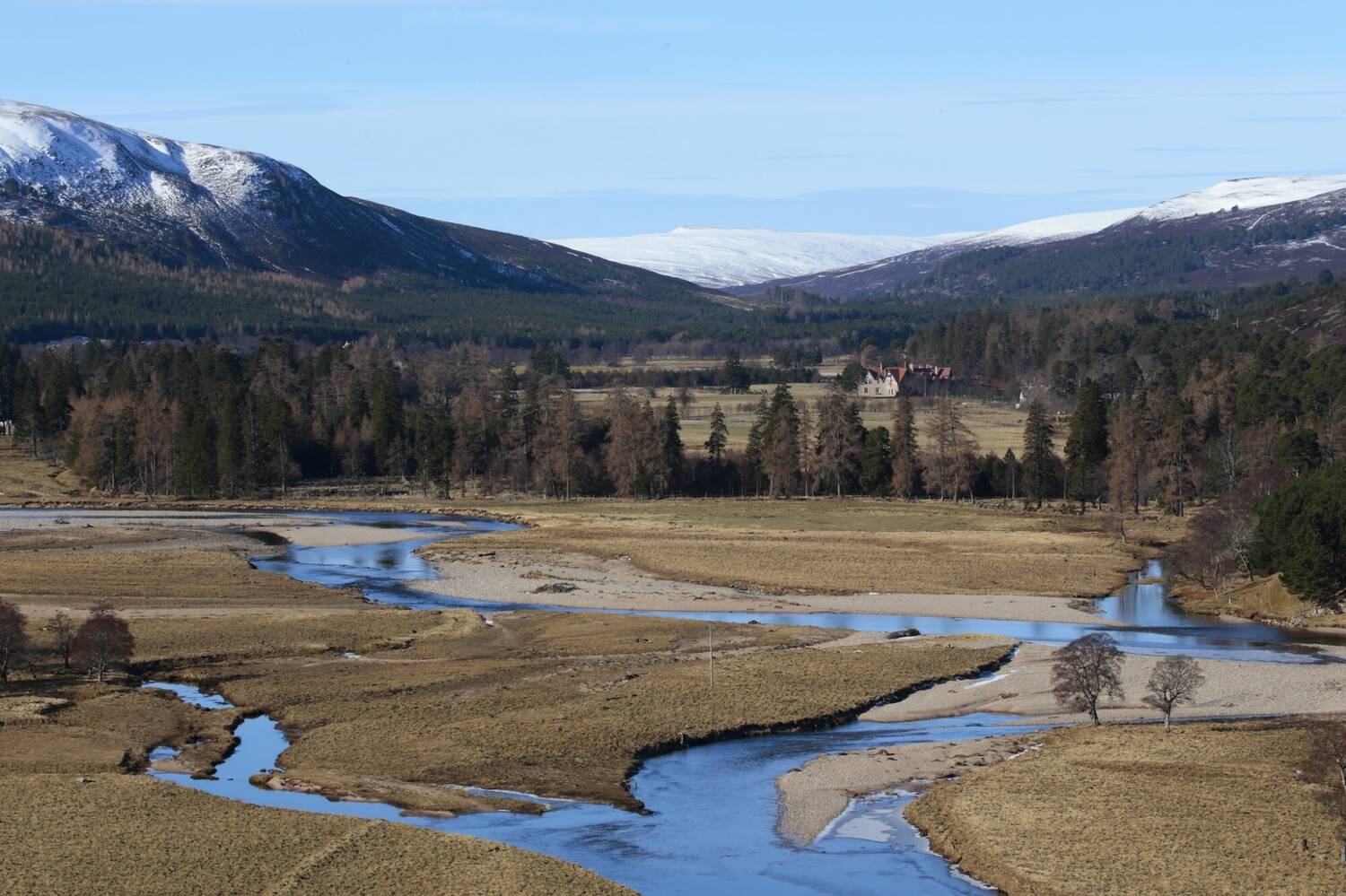 A river meanders through a valley with snow-capped mountains in the background under a blue sky.