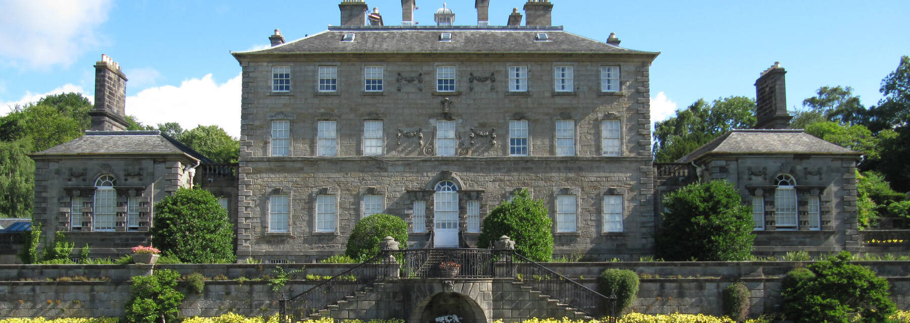 A view of a large, symmetrical Georgian country house. Built in a grey stone, it has a smaller square building each side. Terraced gardens drop down to a lawn in front, with flower beds filled with bright yellow plants.