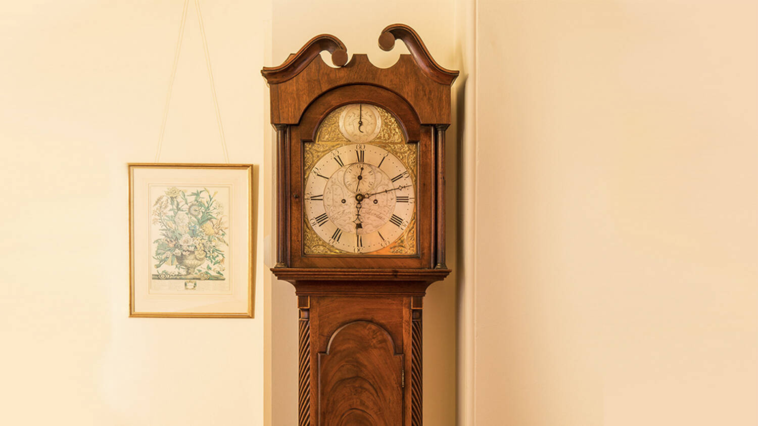 A close-up of the top half of a wooden longcase clock, standing in front of a plain wall next to a framed picture of some flowers.