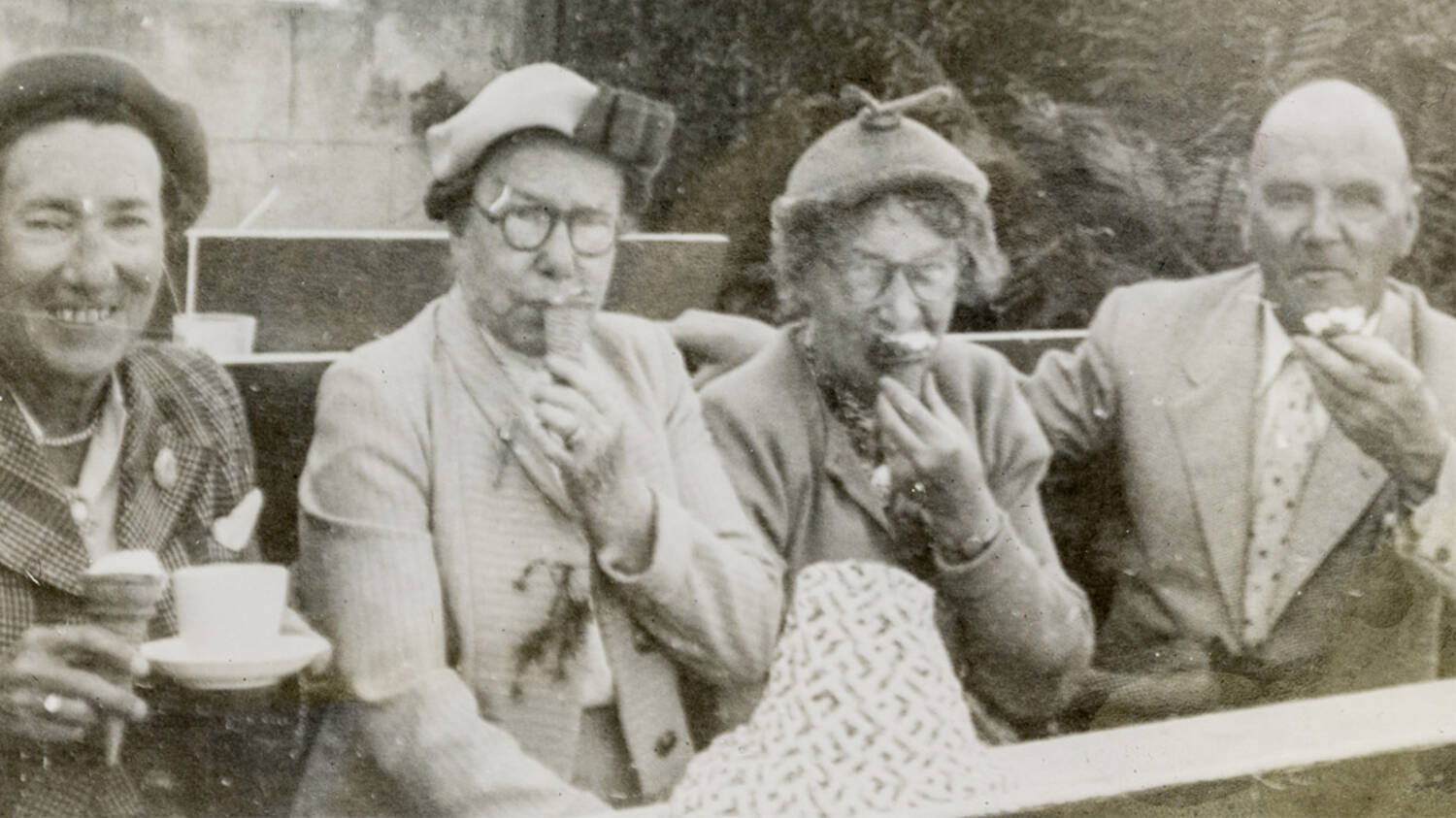 A black and white photograph of three women and a man sitting on a bench outdoors. They are having cups of tea or ice creams (Miss Toward on the far left has both!).