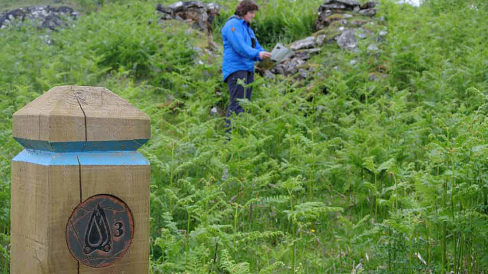 A wooden waymarker post stands in the foreground, with a blue stripe. A lady stands in the background, reading a map.