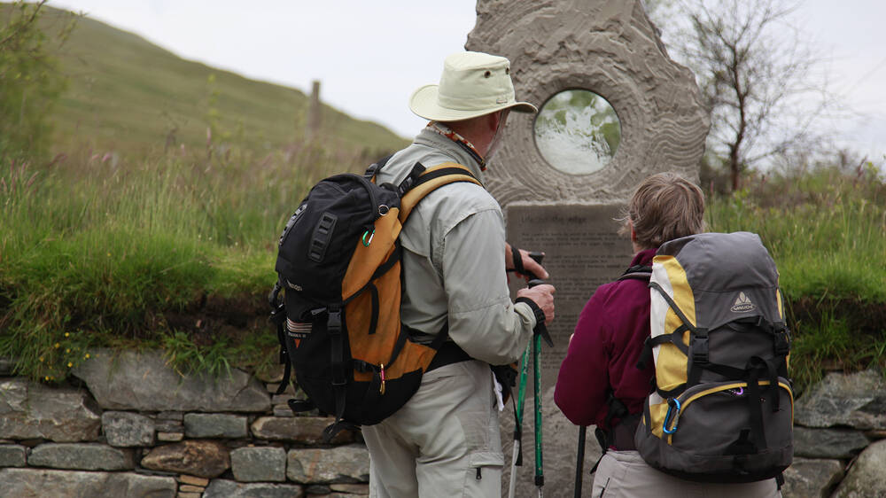 Two walkers wearing backpacks stand in front of a Celtic cross, looking at the inscription.