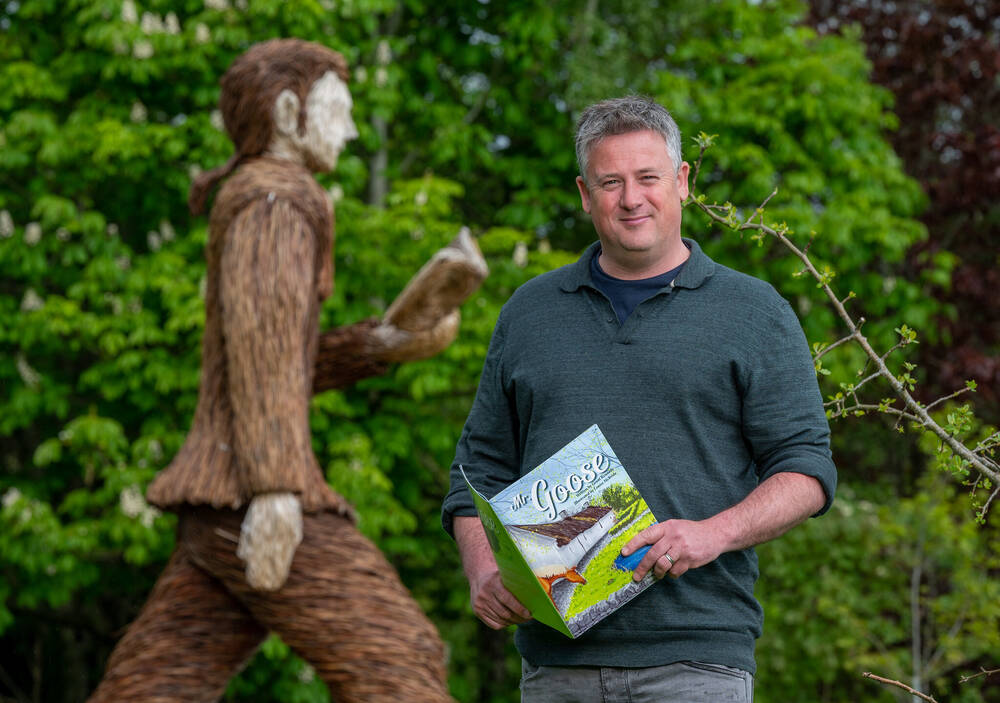 Man in grey jumper holding a book, stands next to large wicker sculpture of a man walking