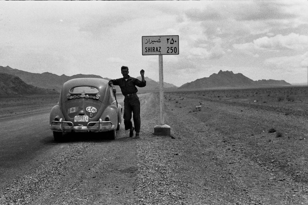 A still from Once More – The story of VIN903847, a black and white film. It shows an old VW Beetle parked at the side of a road in a desert. A man stands beside the car, leaning one arm on its roof.