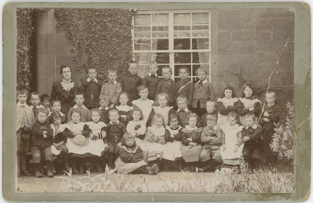A black and white photograph of around 30 young schoolchildren, arranged in rows in front of a stone building with ivy growing up its walls. The windows in the background have lacy curtains. The children are dressed quite formally, with the boys in jackets or kilts, and the girls in white pinafores. Their teacher stands at the back in an austere black dress with a white lace bow.