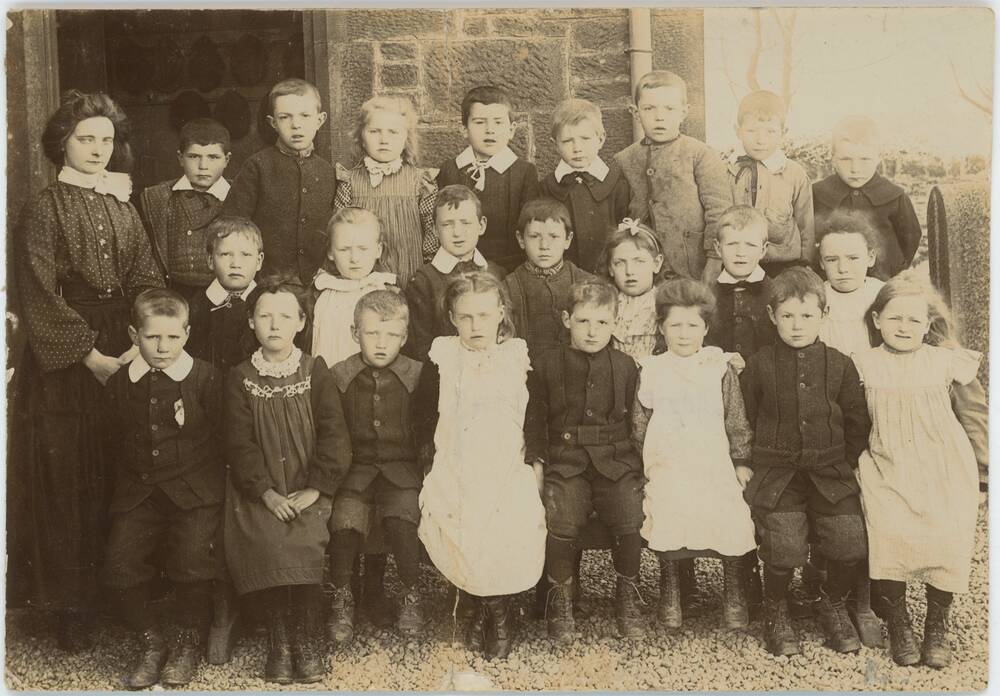 A sepia photograph of a class of 23 young children with their teacher. They stand in three rows outside the school building in a gravelly yard. The boys wear wool jackets and trousers with boots. The girls wear light pinafores over long-sleeved tops. Their young teacher wears a wonderful spotty blouse, with a lace neck and a long black skirt.