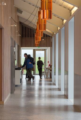A view inside the Bannockburn visitor centre looking down a long light-filled corridor towards a cafe serving area. Several people are at the far end. Unusual orange pendant lights are suspended from the ceiling.