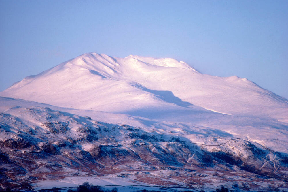 Ben Lawers in winter. The snow-covered mountain stands in the centre of the image, tinged pink from the sun. Snow-covered hills and moorland are in the foreground.