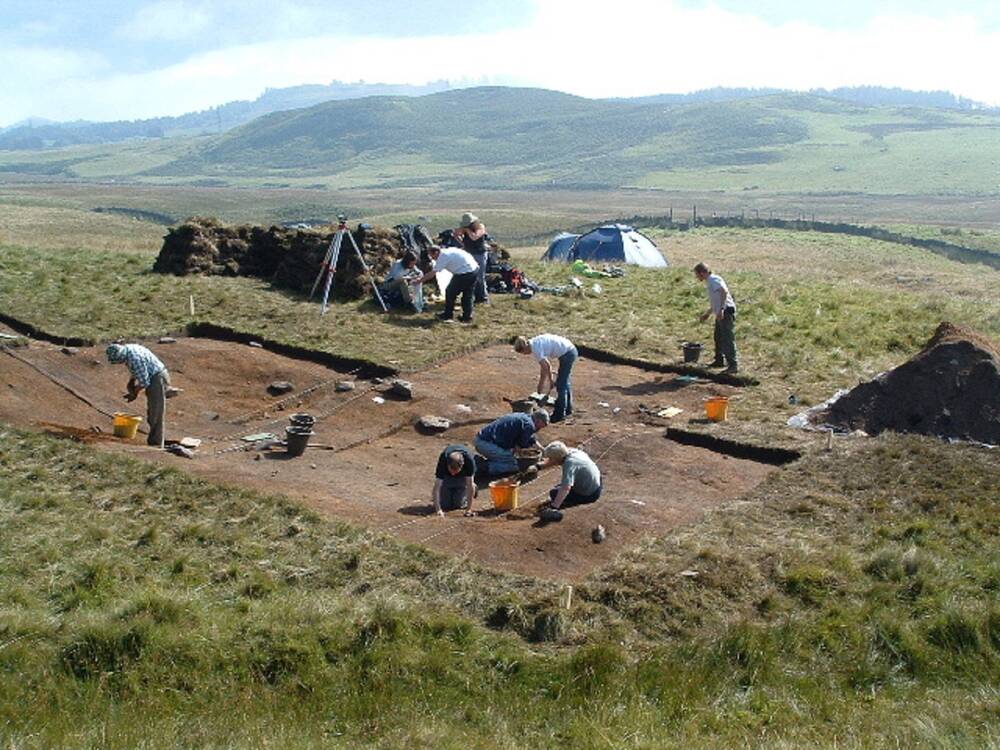 A group of people are at work at an archaeological dig site on a hillside. A square of earth has been dug out, with people kneeling in that recently exposed area. In the background are some ruined stone walls.