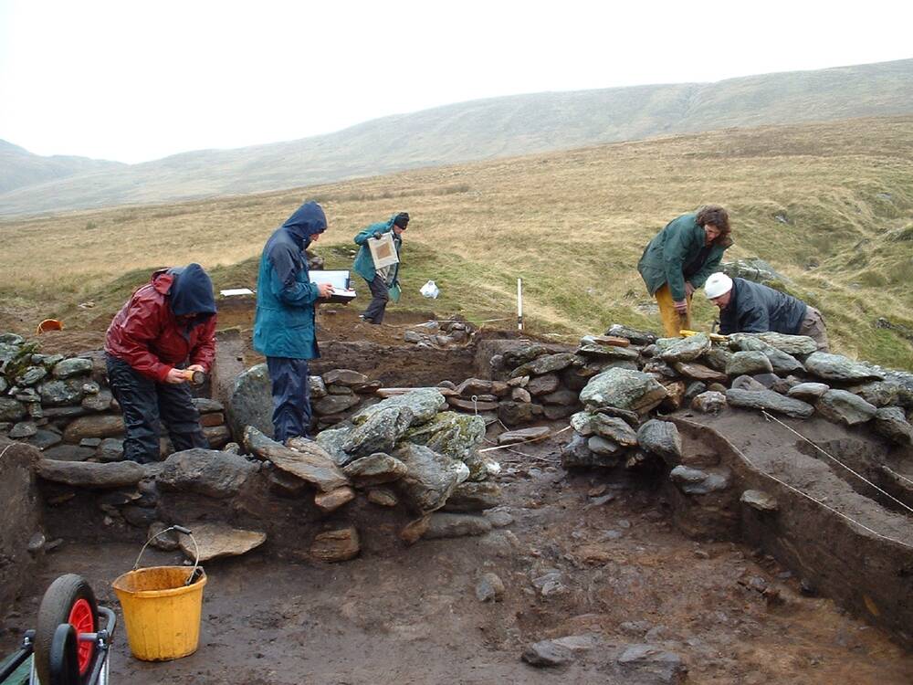 A small group of people are working on some recently exposed stone walls at an archaeological dig on a hillside. The weather is wet, and they are all wearing waterproofs.