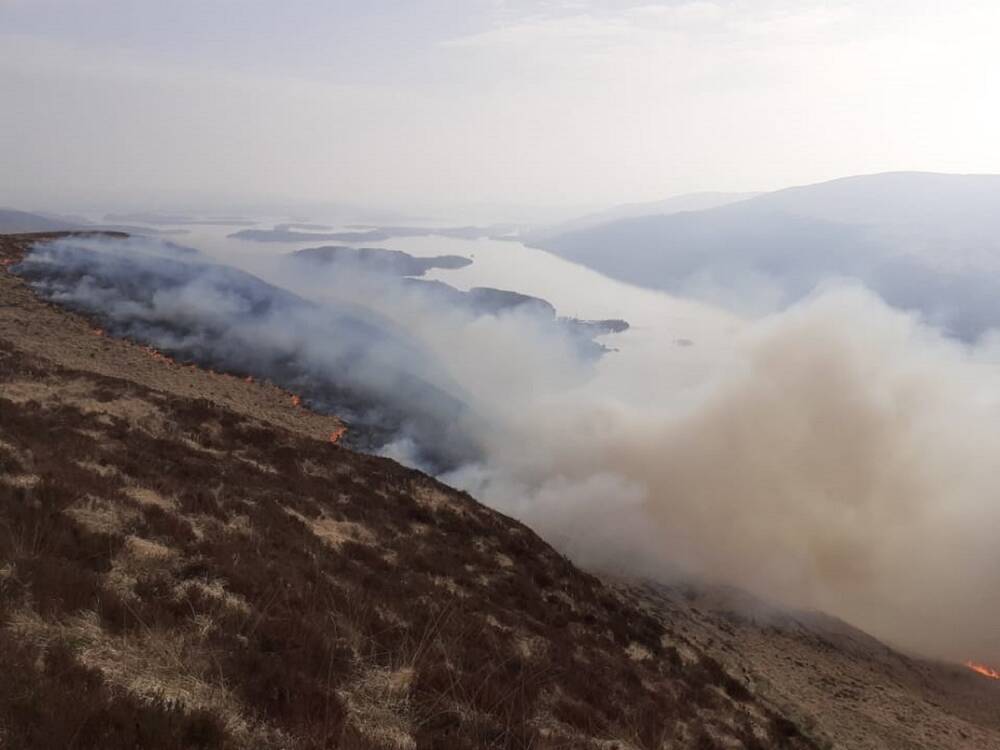 The hillside of Ben Lomond on fire, and a thick cloud of dirty-white smoke obscures the view of the loch below. 