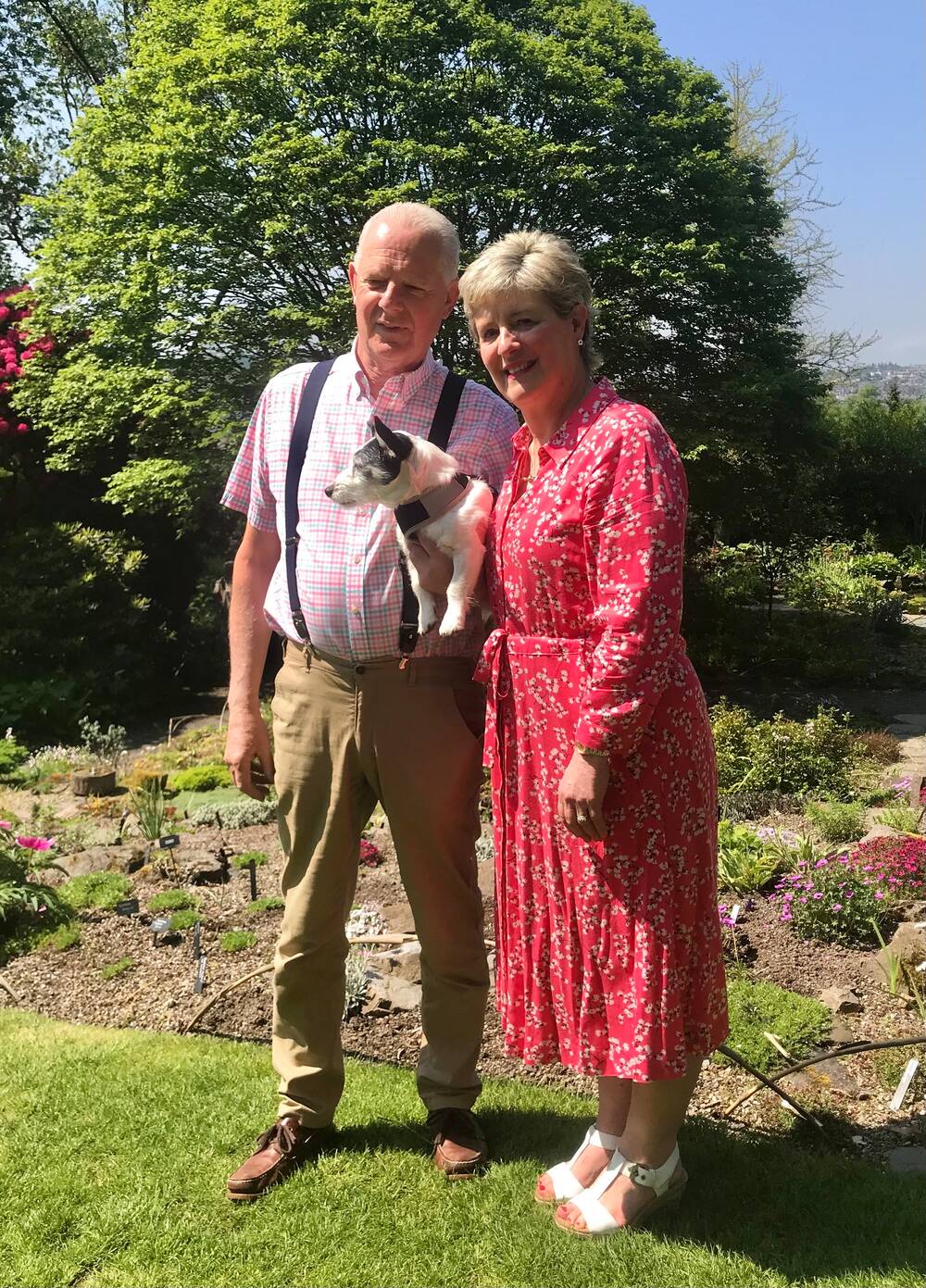 A man and a lady stand beside a rock garden area in Branklyn Garden on a sunny day. They carry a white and black Jack Russell terrier between them.
