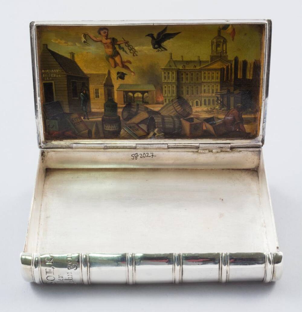 A silver box, shaped like a book, is open, to reveal a tiny painting on the inside of the lid. The box is otherwise empty.