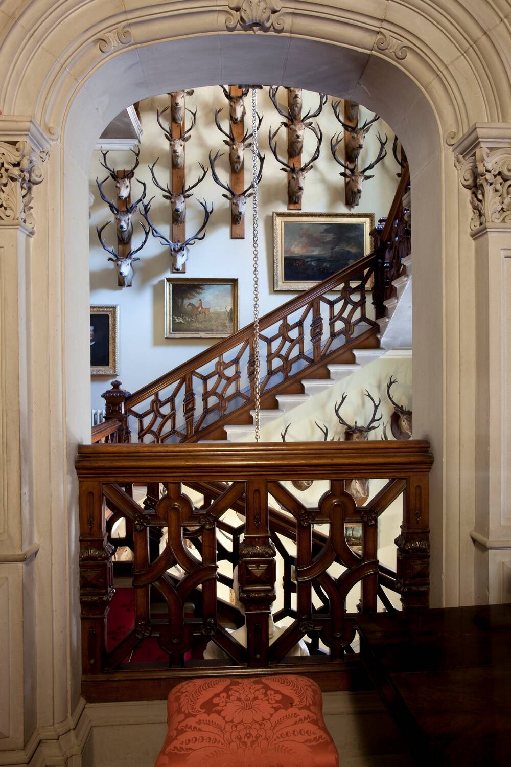 A view of a stairway from a stone arch. The walls of the staircase are covered with mounted stags’ heads, above some framed hunting scenes. The wooden bannister of the stair has a pretty, almost Celtic knot-like carving.